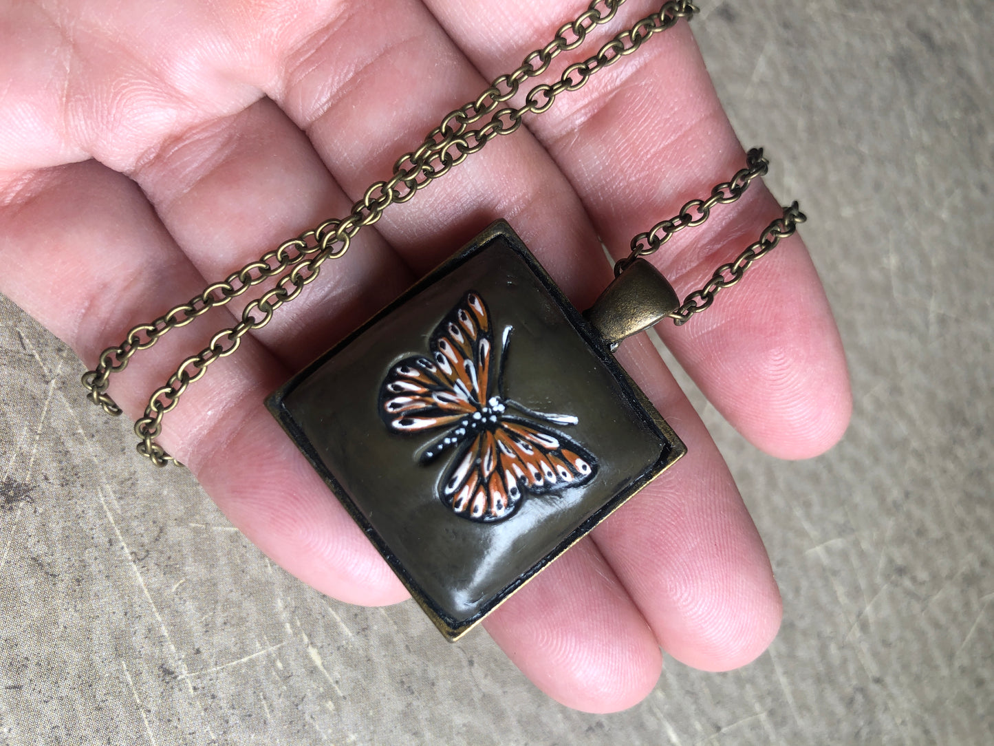 Hand Painted Monarch Style Butterfly on Deep Green Clay Nestled in 1” Square Antiqued Brass Setting Hung with 18” Coordinating Chain